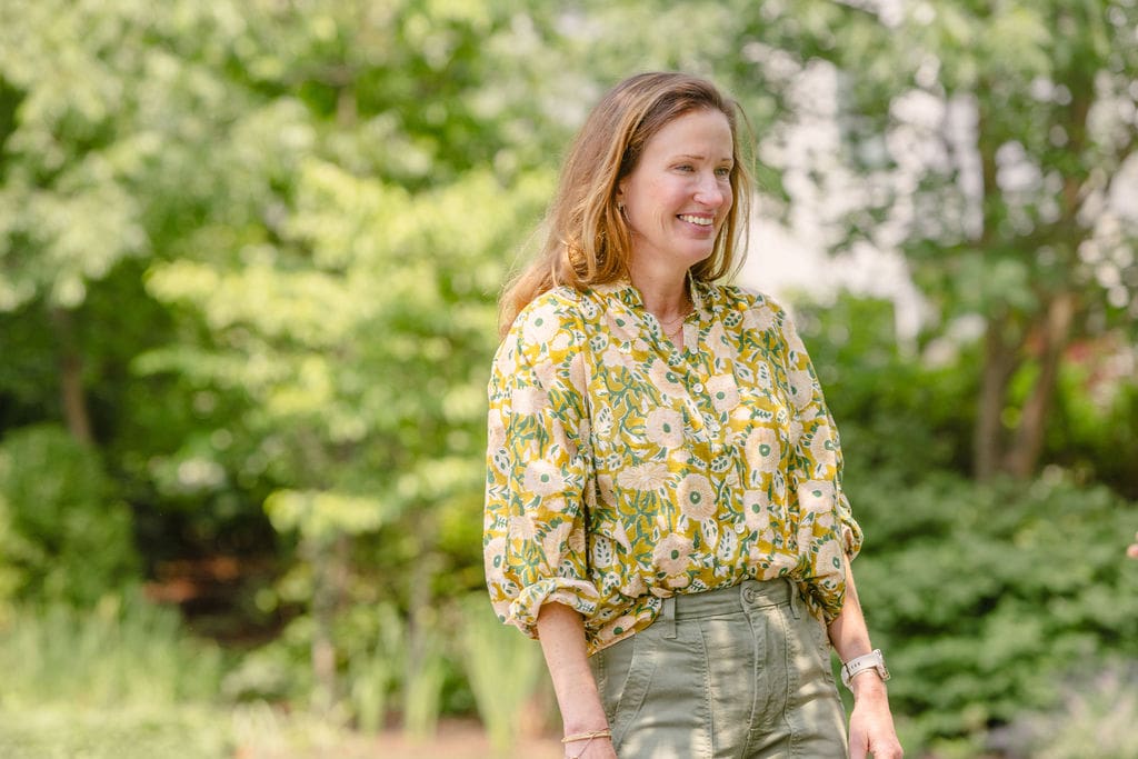 Jenna of Lanier Landscapes wearing floral blouse standing in front of trees